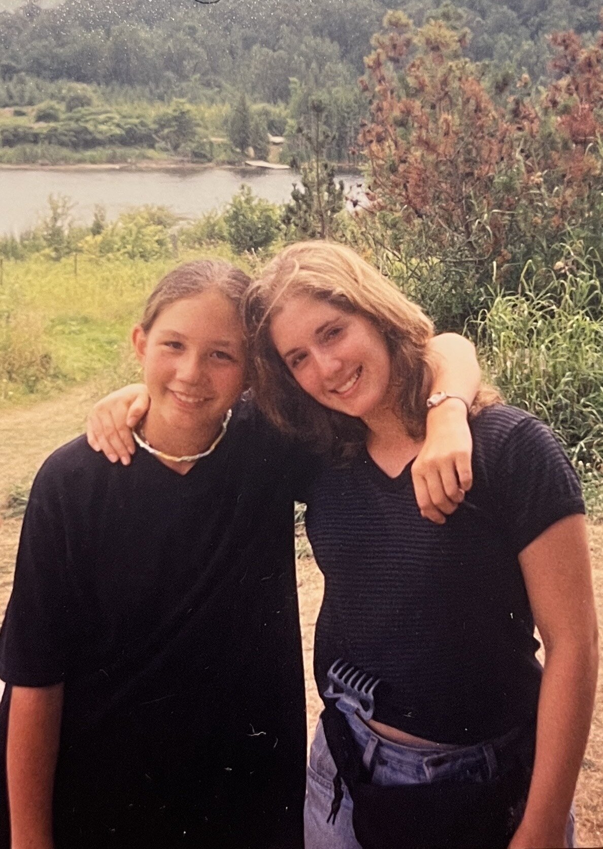 11 year old Sam Wilket stands in front of a lake, smiling with her arm around a blone teenager who is probably her camp counselor.