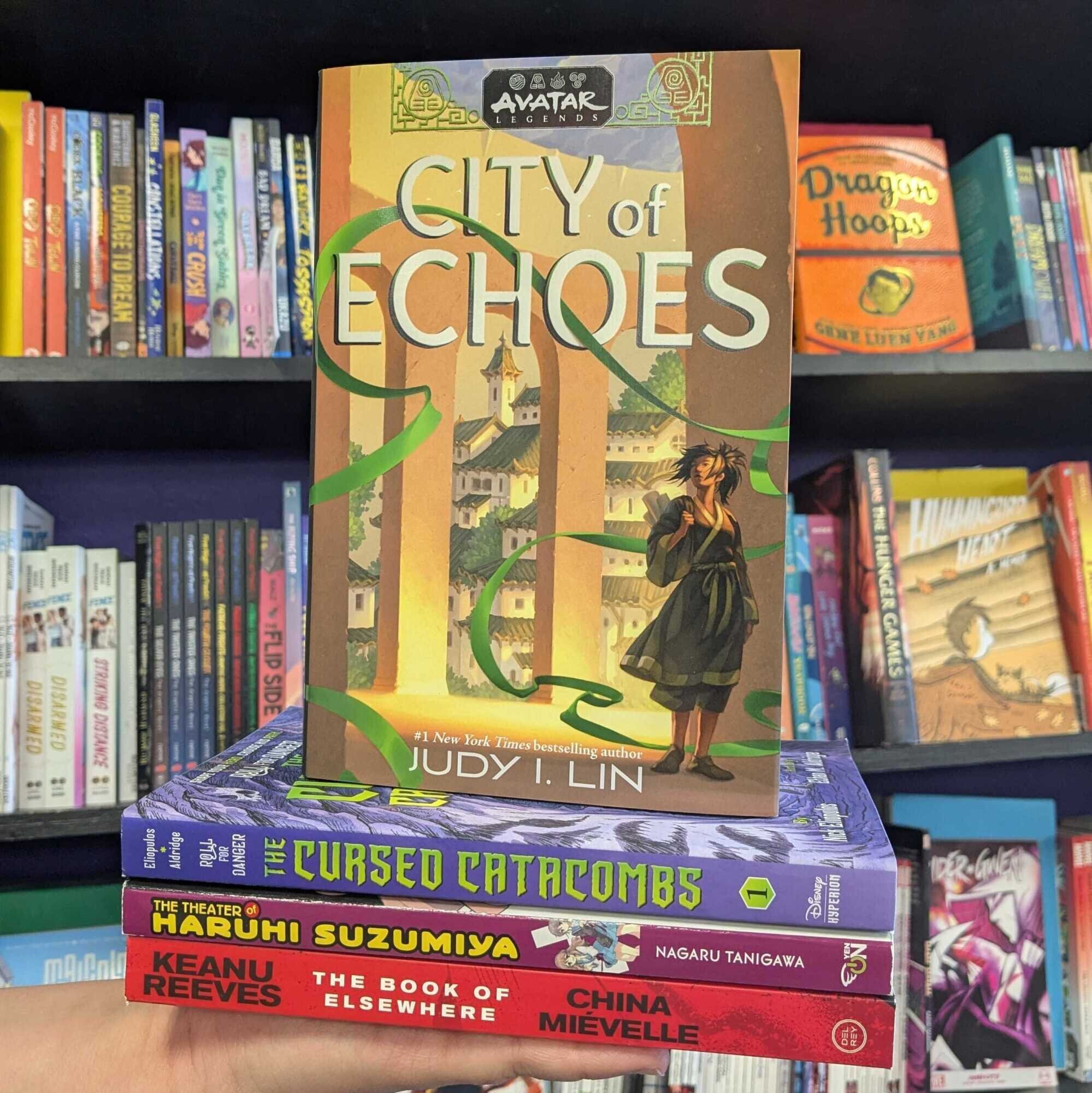 Stack of books. Top to Bottom: "Avatar Legends: City of Echoes," "Roll for Danger: The Cursed Catacombs," "The Theater of Haruhi Suzumiya," and "Book of Elsewhere."
