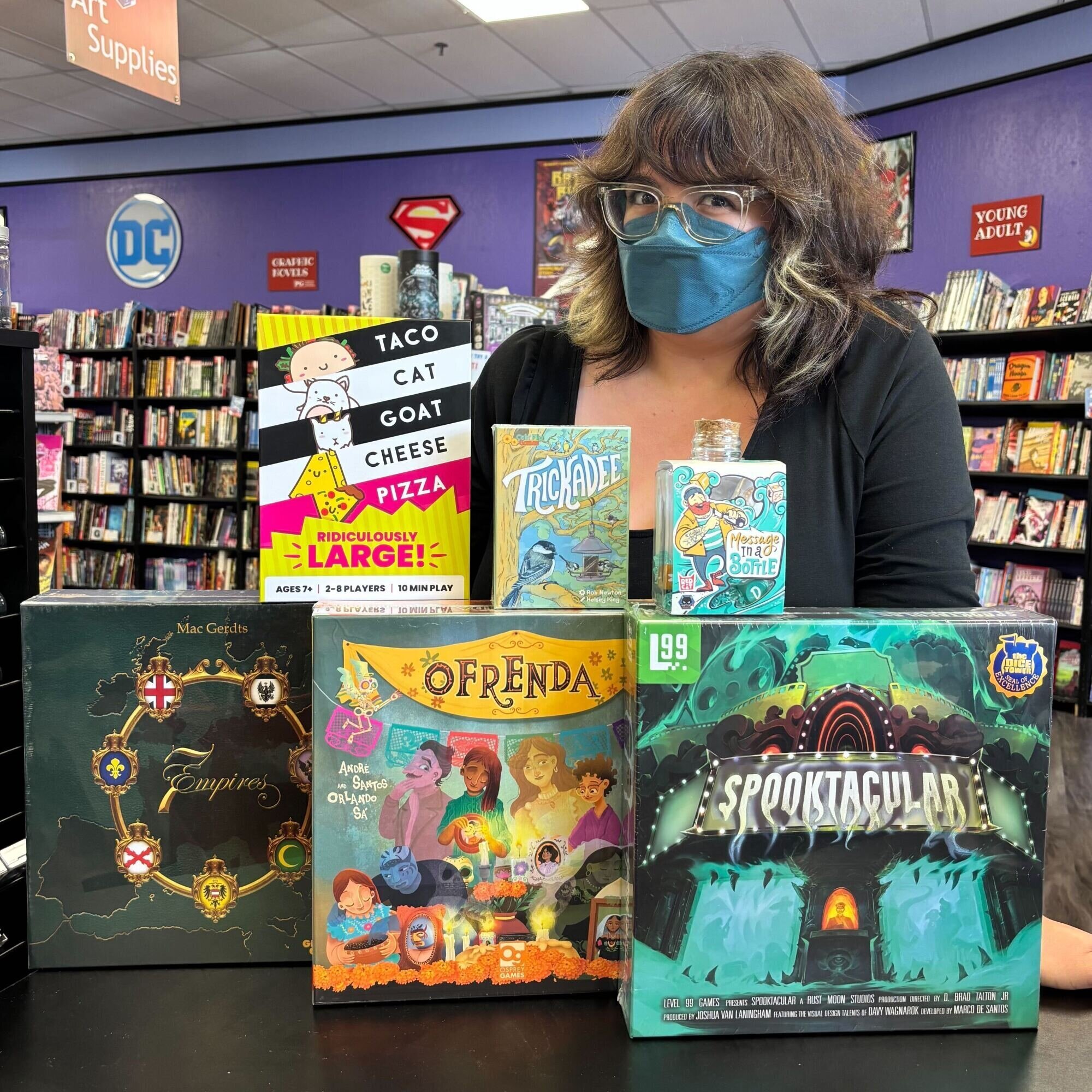 Person posing with display of board games that includes "Taco Cat Goat Cheese Pizza Ridiculously Large!," "Trickadee," "Message in a Bottle," "7 Empires," "Ofrenda," and "Spooktacular."
