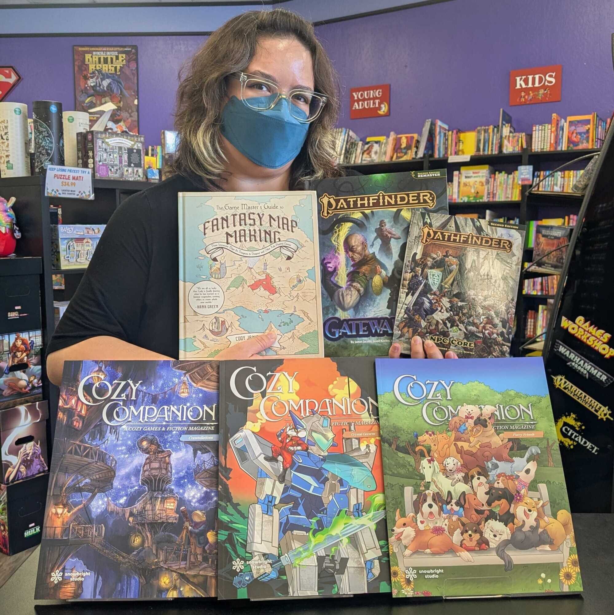 A masked employee stands behind a display of "Cozy Companion" rpg magazines. She holds Pathfinder and Game Masters Guide books.