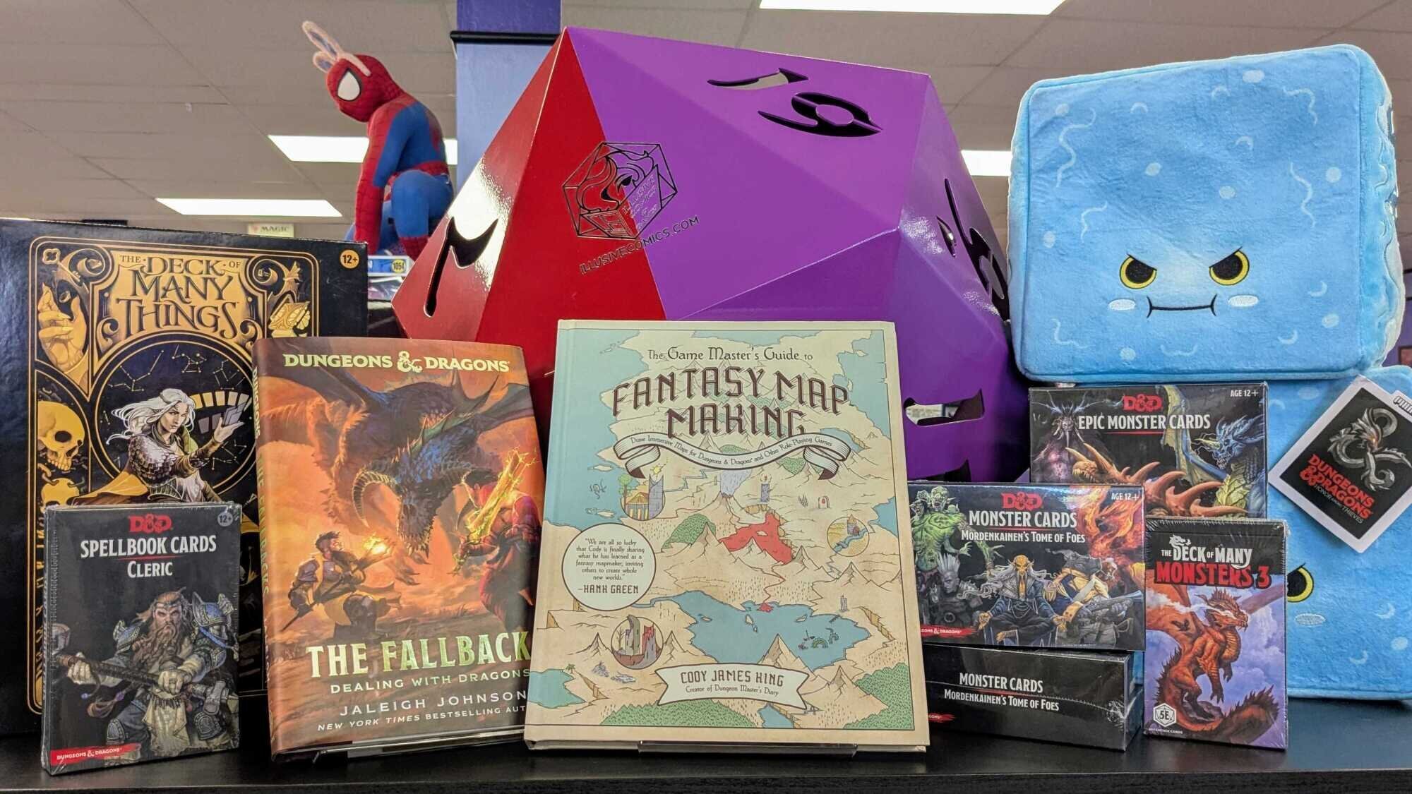A display of Dungeons & Dragons books and accessories. From left to right: The Deck of Many Things boxed set, Cleric Spellbook Cards for 5e, The Fallbacks: Dealing with Dragons novel, The Game Master's Guide to Mapmaking, monster card sets, and gelatinous cube plushies.
