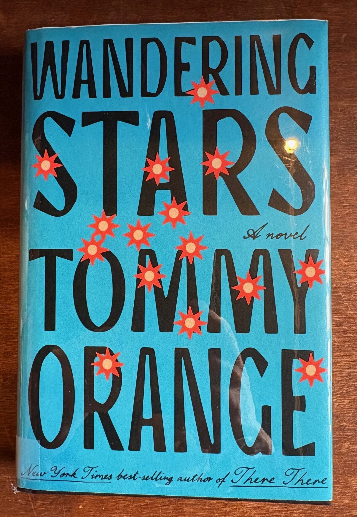 A hardcover copy of WANDERING STARS by Tommy Orange sits on a brown table. The book is wrapped in a dust jacket