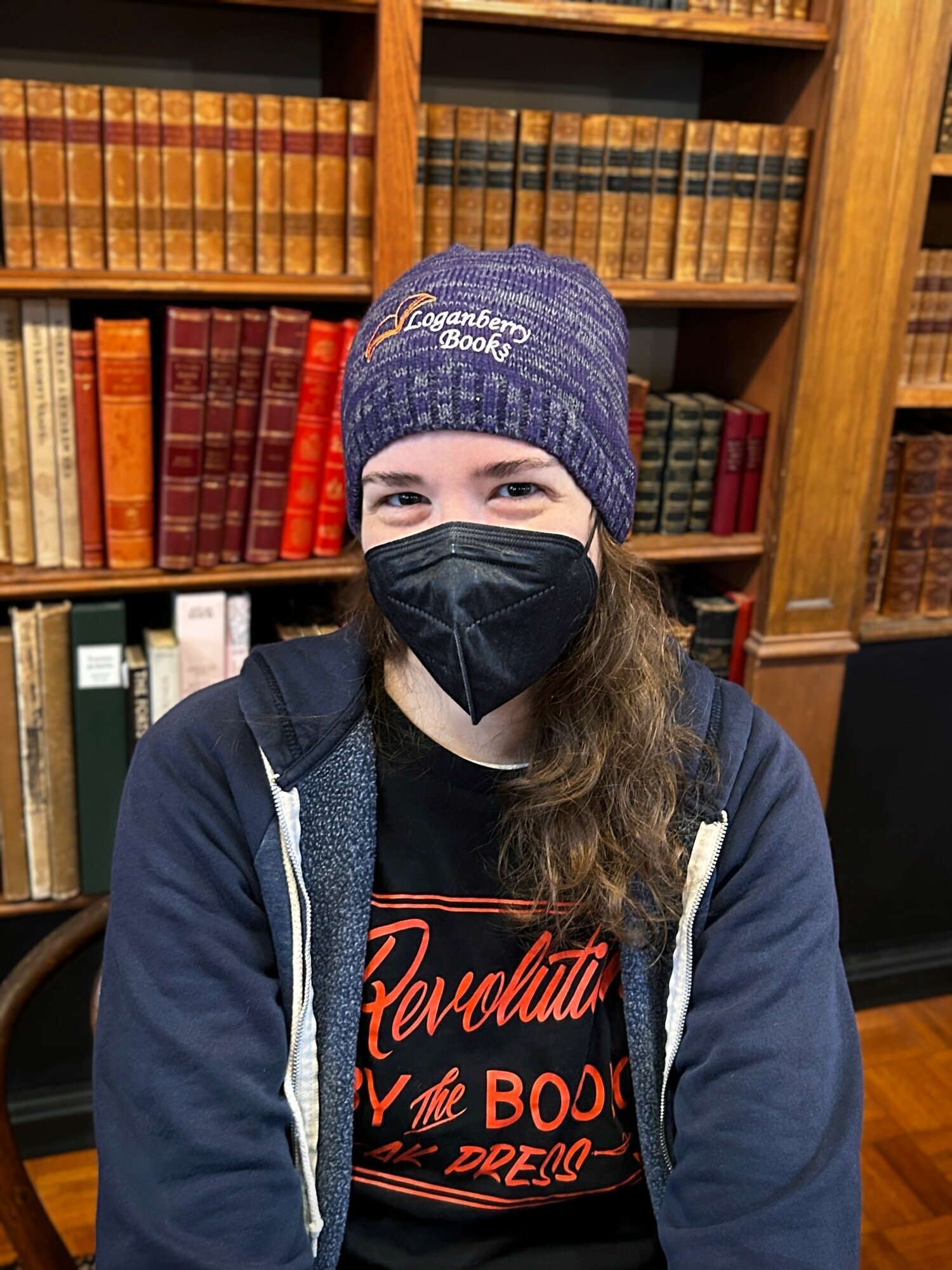 Woman wearing a mask models a purple beanie in front of a bookshelf