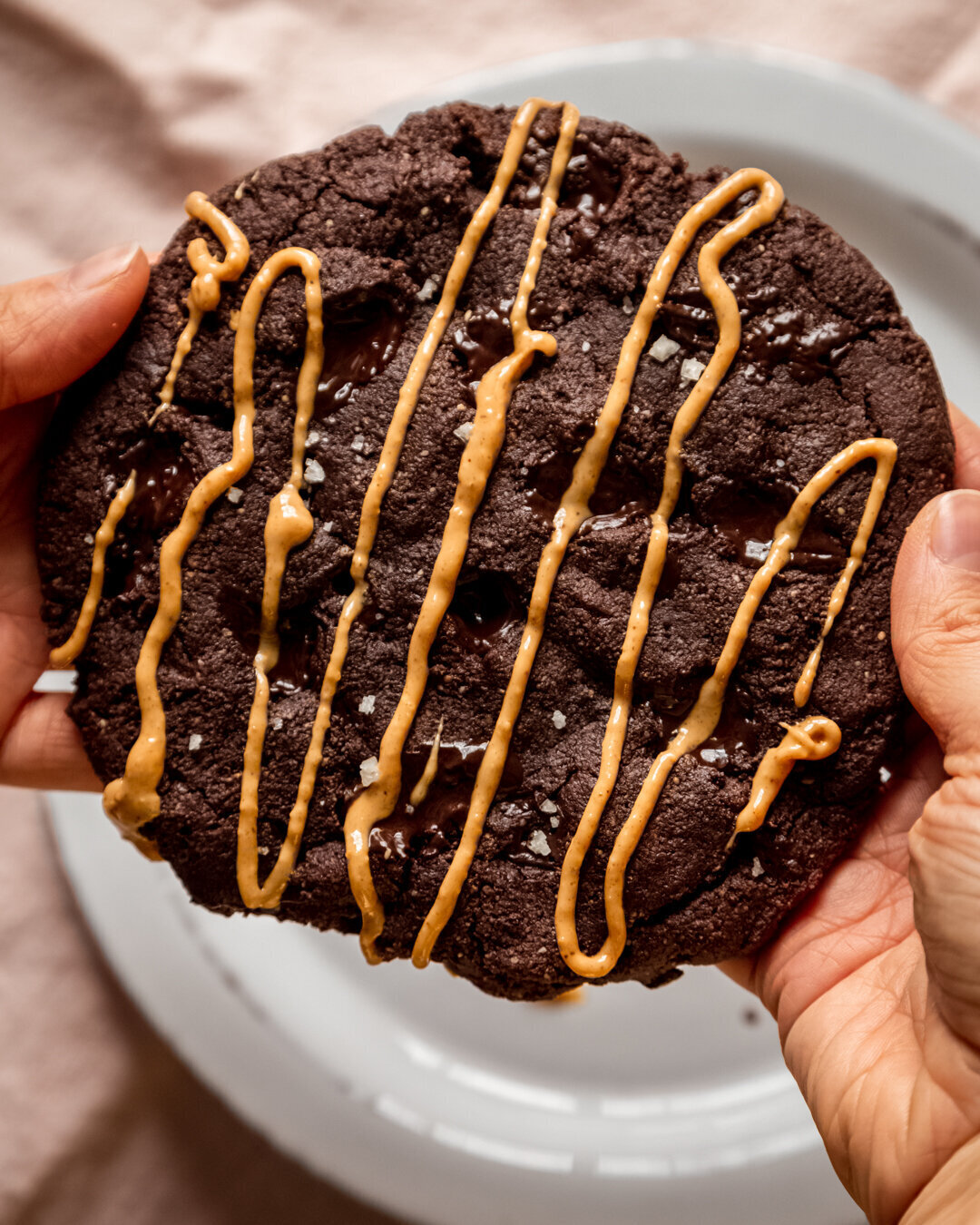 An overhead image shows a set of hands holding a jumbo double chocolate cookie drizzled with peanut butter.