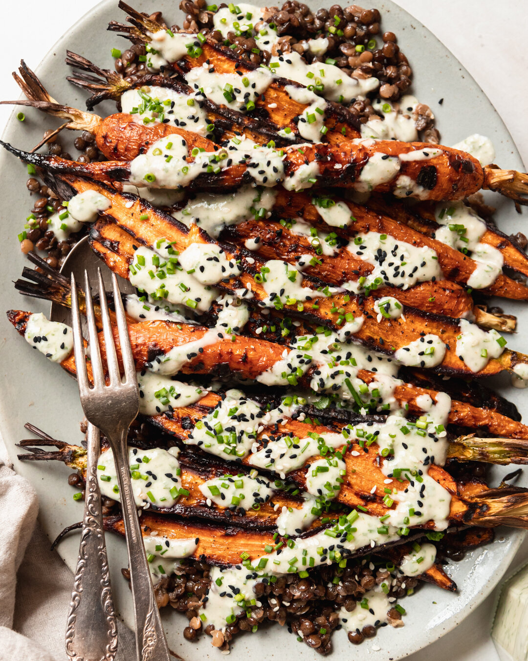 An up close, overhead shot shows a light blue platter topped with cooked french lentils and grilled whole carrots, all topped with a creamy chive-flecked sauce. Extra chives and sesame seeds garnish the dish.
